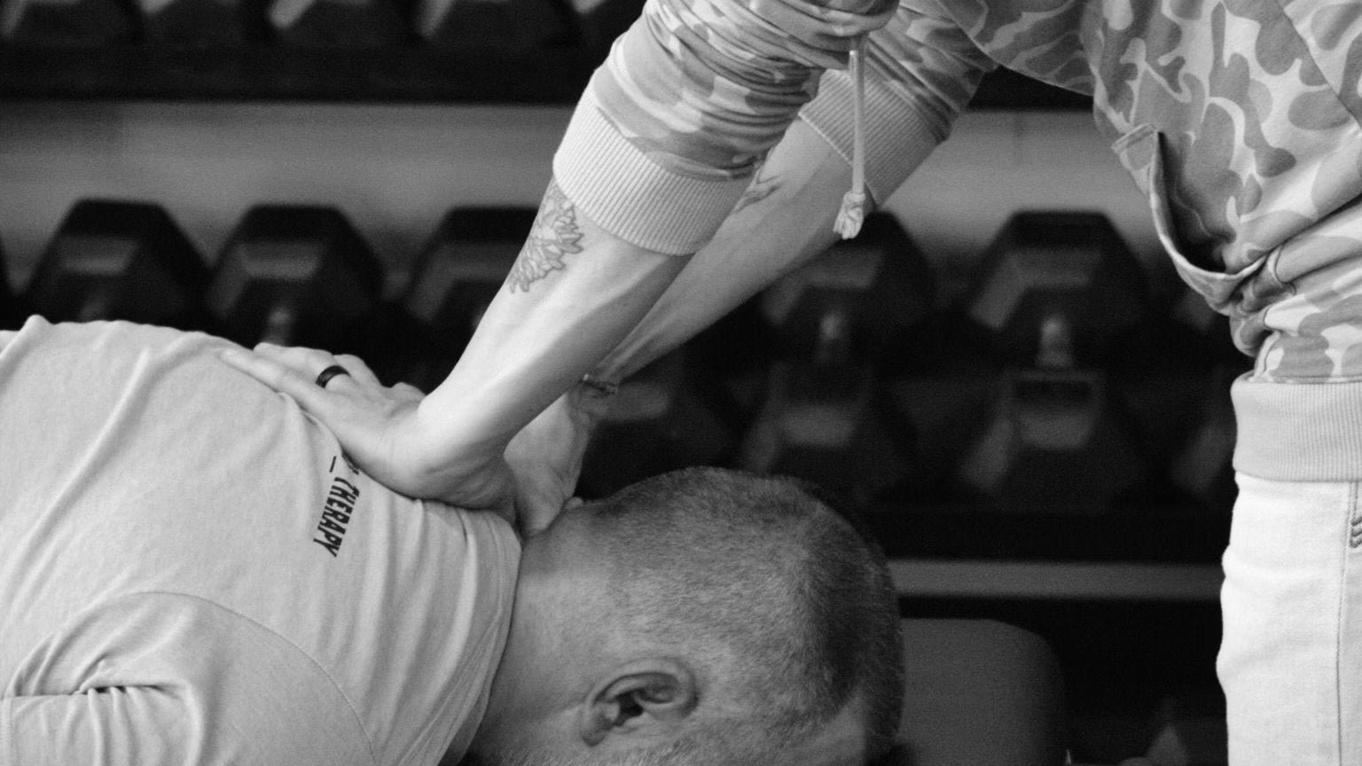 Person performing a massage on another person in a gym setting with weights in the background.